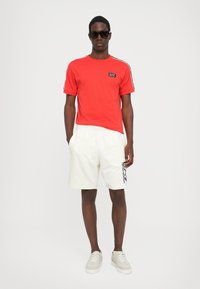 Red t-shirt with black logo, white logo-printed shorts, and beige sneakers. The shirt has short sleeves and a crew neck design.