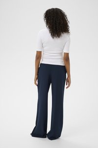 Sieviete ar cirtainiem matiem, tērpusies baltā īsroku T-kreklā un brīvā tumši zilā biksēs, stāvot ar muguru pret vienkāršu fona.