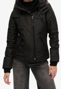 Femme portant une veste noire zippée avec plusieurs poches zippées et un jean gris foncé, debout les mains détendues le long du corps.
