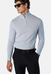 Maglione zip-up blu chiaro con maniche lunghe, texture morbida e colletto alto, abbinato a pantaloni scuri e una cintura nera intrecciata con un accento marrone.
