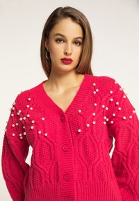 Cardigan tricotat roz aprins, cu un model de cablu, închidere cu nasturi și accente de perlă pe umeri. Croi texturat, supradimensionat.