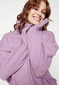 Pull en maille violet oversized avec un col montant, doté de manches amples et d'une texture douce, drapé sur les épaules du modèle.