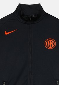 Melns jaku ar rāvējslēdzēju, kas izgatavota no viegla auduma. Priekšpusē ir oranžs Nike logo kreisajā krūtī un oranžs apļa emblem.
