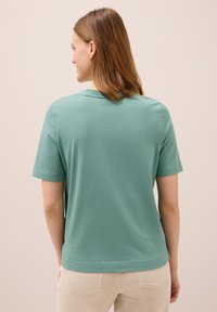 Mujer con cabello castaño claro hasta los hombros, vistiendo una camiseta verde de manga corta y pantalones beiges, de espaldas contra un fondo liso.