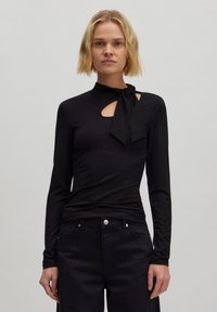 Schwarzer Langarm-Top mit hohem Kragen und Bindungsdetail, das einen Ausschnitt auf der Brust hat. Der Stoff ist glatt und eng anliegend.