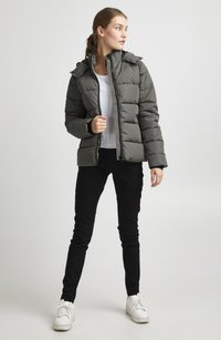 Šedá péřová bunda s kapucí, prošívaný design, přední zip a přiléhavý střih, spárována s černými kalhotami a bílými teniskami.