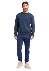 Dunkelblaues Sweatshirt mit ELB-Logo, gerippte Bündchen und Saum, kombiniert mit passenden Sweatpants. Die Schuhe sind hellbeige mit dunkleren Akzenten.