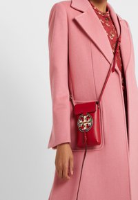 Rød lædertaske med en cirkulær metal-logo detalje, kombineret med en lyserød uldfrakke. Tasken har en lang rem og et lukket klapdesign.