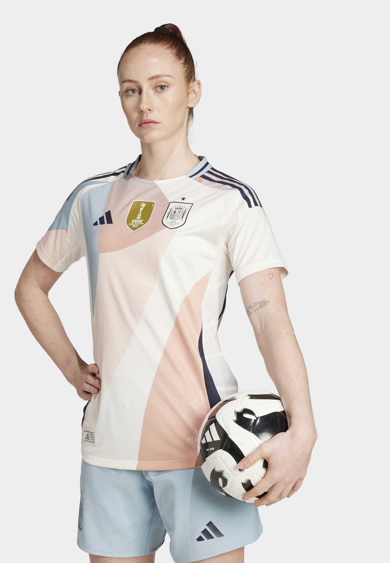 Camiseta de fútbol en blanco con patrones geométricos en rosa y azul, marca Adidas, logo de la FIFA y pantalones cortos a juego en azul claro, sosteniendo un balón de fútbol en blanco y negro.