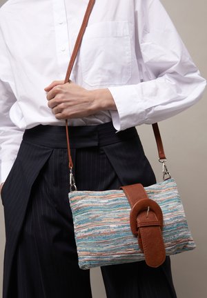 Oseba, ki nosi belo majico in črne hlače s črtami, drži večbarvno tkano torbico čez ramo z rjavim usnjenim trakom in zaponko.