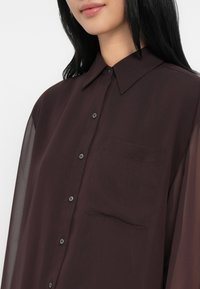 Mørk burgunder knappskjorte med krage, gjennomsiktige lange ermer, en brystlomme og matchende knapper. Jevn tekstur, lett stoff.