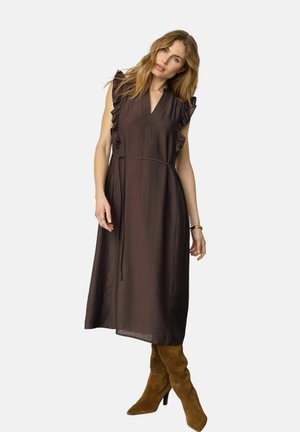 Kvinde iført en ærmeløs mørkebrun midi-kjole med flæser på skuldrene og et tyndt bælte, kombineret med brune ruskindsstøvler til knæet.