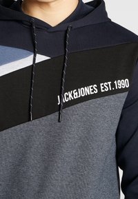 Huva-sweatshirt i marinblå, svart och grå nyanser. Har en geometrisk färgblockdesign och vit logotext. Texturerat tyg, justerbara snören.
