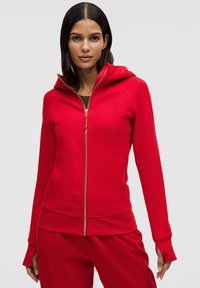 Červená mikina na zip z měkké látky s předním zipem, kapucí a bočními kapsami; má otvory na palce v manžetách a těsný střih.