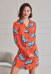 Woman with short brown hair wearing a bright orange buttoned dress with blue bird prints, standing indoors near a light gray textured chair.