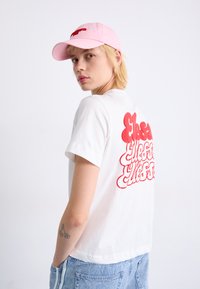 Hvit bomulls t-skjorte med stor rød bobletekst på baksiden. Modellen har på seg en rosa caps og lyseblå denimjeans.