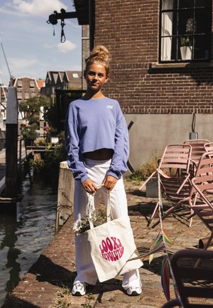 Young woman in cropped purple sweatshirt and white pants holding a tote bag with flowers near canal and brick building on sunny day.