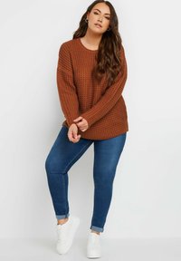 Brązowy sweter dzianinowy z teksturowanym wzorem, luźny krój, okrągły dekolt, zestawiony z ciemnoniebieskimi dżinsami i białymi sneakersami.
