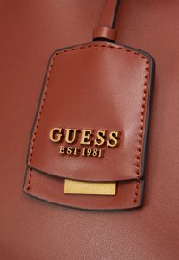 Sac à main en similicuir marron avec une étiquette logo dorée. Texture lisse, accents cousus et forme rectangulaire. Détails en métal doré.