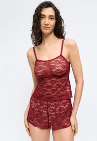 Femme portant un caraco en dentelle bordeaux et un short assorti, debout contre un fond clair uni, faisant face à l'avant avec une main baissée.