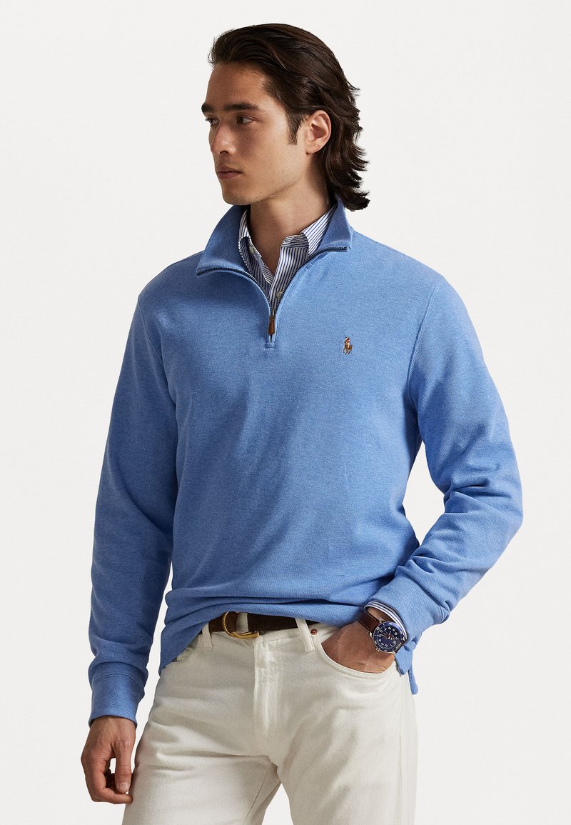 Sweater pullover azul claro com gola com fecho, tecido texturizado e pequeno logotipo em destaque. Usado sobre uma camisa de colarinho riscada e combinado com cal�ças brancas.