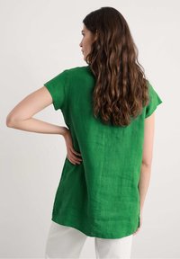 Tunic in lino verde con maniche corte, spacchi laterali e una vestibilità rilassata, caratterizzata da un tessuto testurizzato e un orlo arrotondato, abbinata a pantaloni bianchi.