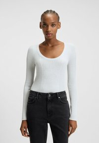 Body a maniche lunghe a coste grigio chiaro con scollo rotondo, abbinato a jeans neri a vita alta con tasche frontali e bottone visibile.