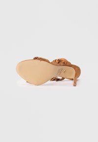 Tan suede high-heeled sandal with an open toe, detailed with twisted fabric accents, a textured sole, and a slender stiletto heel.