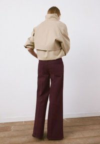 Osoba o jasnobrązowych włosach, ubrana w beżową krótka kurtkę i szerokie ciemnofioletowe spodnie z wysokim stanem, stojąca na drewnianej podłodze.