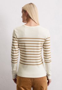 Vrouw met lang blond haar, gekleed in een crèmekleurige shirt met lange mouwen en horizontale lichtbruin strepen, en een bruine broek, staande voor een gestructureerde witte muur.
