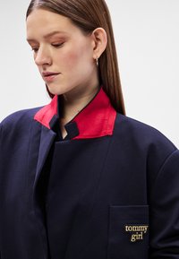 Ναυτικό σακάκι με κόκκινο γιακά, διαθέτει τσέπη στο στήθος με το «tommy girl» κεντημένο σε χρυσό. Λείο υφή, μοντέρνος σχεδιασμός.