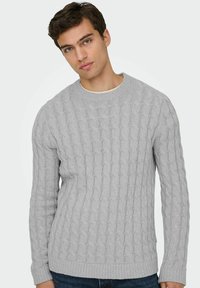 Jeune homme portant un pull en maille torsadée gris clair et un jean foncé, se tenant devant un fond blanc uni.