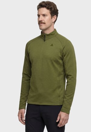 Mann trägt einen olivgrünen Pullover mit Viertel-Reißverschluss und langen Ärmeln sowie schwarze Hose, steht vor einem schlichten hellen Hintergrund.