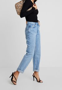 Lichtblauwe, hooggetailleerde spijkerbroek met opgerolde boorden, gecombineerd met een zwart, geribd, lang mouwen top en zwarte stiletto sandalen, geaccentueerd met een tas met patroon.