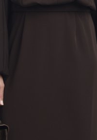 Črna obleka z dolgimi, napihnjenimi rokavi in gladko teksturo. Roka drži črno torbico, ki prikazuje čiste linije in eleganten dizajn.