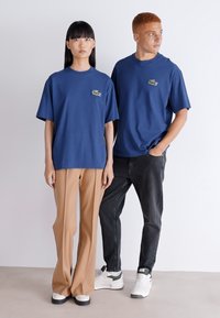 Camisetas de algodón azul de cuello redondo con acento de logo, combinadas con pantalones beige y jeans oscuros. El calzado incluye zapatillas blancas con detalle en verde.