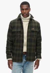 Camisa de franela en verde oscuro con patrón de cuadros negros, con botones en la parte delantera, dos bolsillos en el pecho, usada sobre un suéter de cuello alto crema y combinada con jeans desgastados.