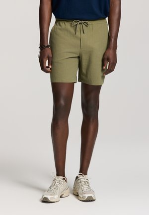 Mann trägt olivgrüne Shorts mit Kordelzug und beige Laufschuhe, steht vor einem schlichten hellen Hintergrund.