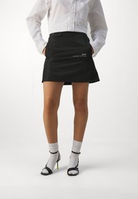 Jupe noire avec poches, texture lisse, coupe ajustée, bouton à la taille. Associée à des chaussettes blanches et des sandales à talons noires avec des accents verts.