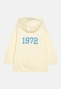 Dzeltena kapuce ar lielu zilu "1972" uzdrukājumu aizmugurē un kontrastējošu Nike logo. Iespējama ar šnurēšanas kapuci un ribotām aprocēm.