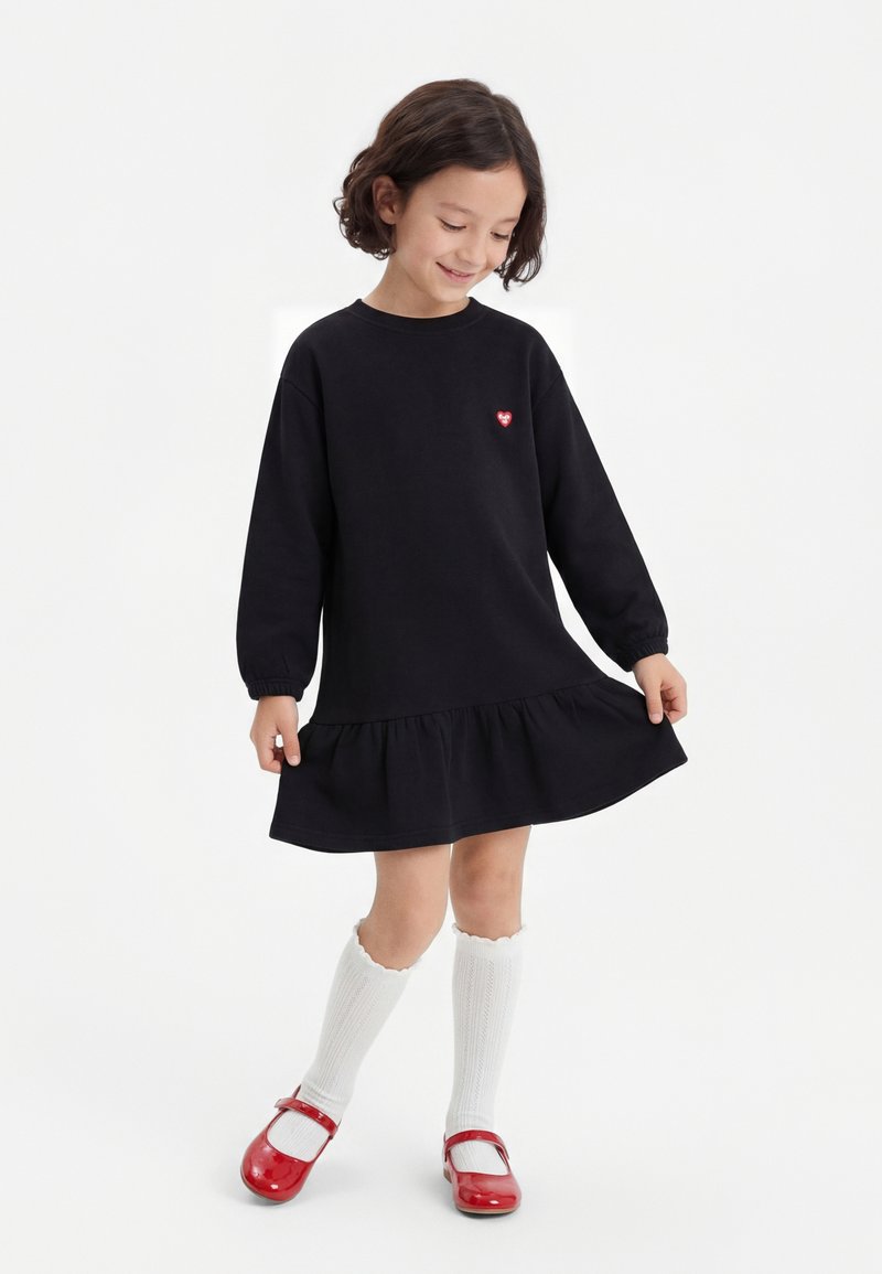 Nuori tyttö pukeutuneena mustaan pitkähihaiseen paitamekkoon, jossa on röyhelöhelma, valkoiset polvisukat ja punaiset Mary Jane -kengät, hymyillen ja pitelemässä mekon sivuja.