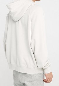 Ljusgrå hoodie gjord av mjukt tyg, med dragsko i huvan, ribbade muddarna och en avslappnad passform. Bak sidan visar minimal detaljering.