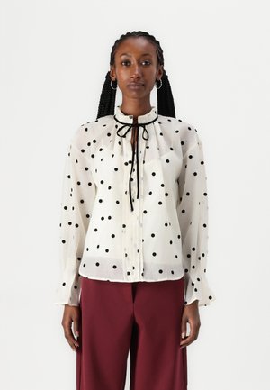 Blusa blanca con lunares negros, cuello alto, corbata negra en el cuello y mangas largas, hecha de tela transparente. Combinada con pantalones burdeos.