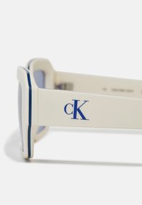Bele sončne očala z modrim trakcem, z debelim okvirjem in izstopajočim modrim logotipom "cK" na roki. Leče imajo rahlo toniranje.