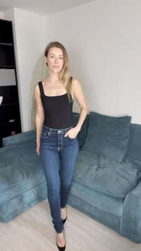 Bodysuit negro sin mangas combinado con jeans azules de talle alto. La modelo está de pie sobre un suelo claro al lado de un sofá de pana en tono teal.