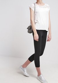 Camiseta blanca con detalle de encaje en las mangas, combinada con leggins negros y zapatillas plateadas; el modelo posa frente a un fondo claro.