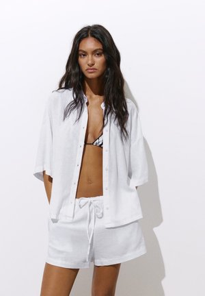 Femme aux longs cheveux foncés portant une chemise blanche ouverte à boutons, un short blanc à cordon de serrage et un haut de bikini noir et blanc.