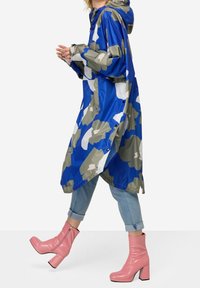 Blå och grå kamouflage-mönstrad regnjacka med huva, glänsande textur och tryckknappar, tillsammans med knähöga rosa klackstövlar.