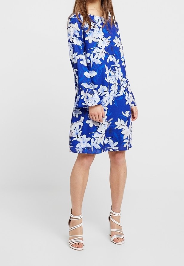Femme portant une robe bleue à imprimé floral blanc et manches longues, associée à des sandales blanches à talons hauts et à brides.