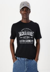 Black cotton T-shirt with white printed logo and text. Worn with denim shorts. Gray cap featuring colorful embellishments.